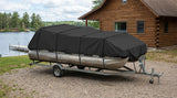 A black boat cover secured over a pontoon boat on a trailer, parked outdoors on a gravel surface in front of a wooden cabin with a lake and trees in the background.